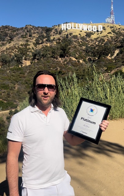 A man in sunglasses holds a 'Platinum' certificate outdoors with the Hollywood sign visible in the background.